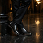 Black leather Chelsea boots on a polished dark stone floor reflecting chandelier light, black trouser hem breaking over the boot