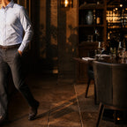 Man in a light blue shirt and charcoal trousers standing in a warmly lit restaurant at dusk, cropped by the left edge of the frame