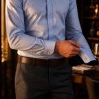 Close crop of a hand adjusting the buttoned cuff of a light blue shirt above charcoal trousers and a brown leather belt