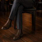 Seated man from the knee down: dark brown Chelsea boots on a dark wooden floor, charcoal trouser hem breaking over the boot