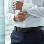 Close crop of hands fastening the cuff button of a white shirt above navy trousers and a black leather belt with a matte square buckle