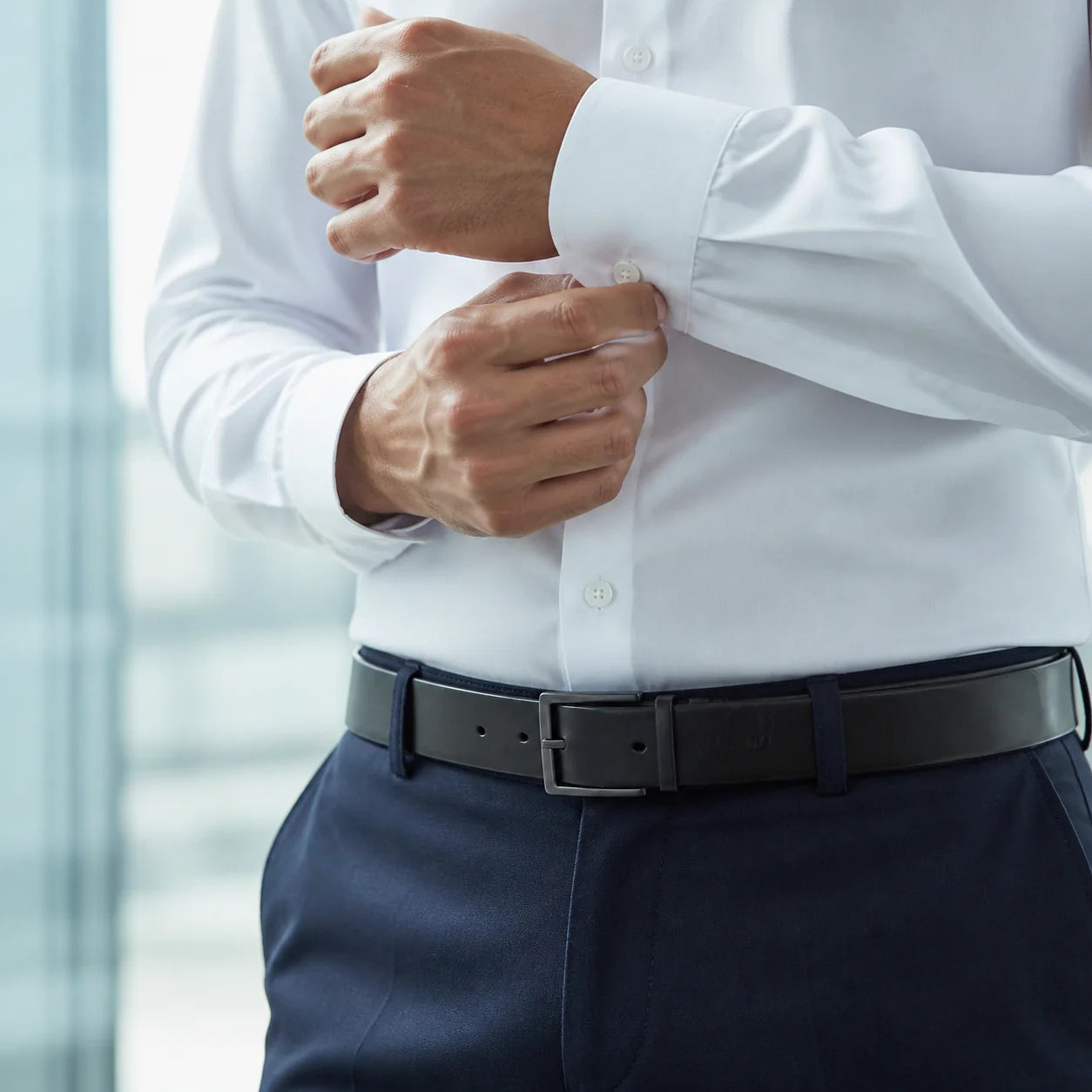 Close crop of hands fastening the cuff button of a white shirt above navy trousers and a black leather belt with a matte square buckle