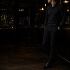 Man in an all-black shirt and trousers standing at a dimly lit bar, low three-quarter angle, face turned away from camera