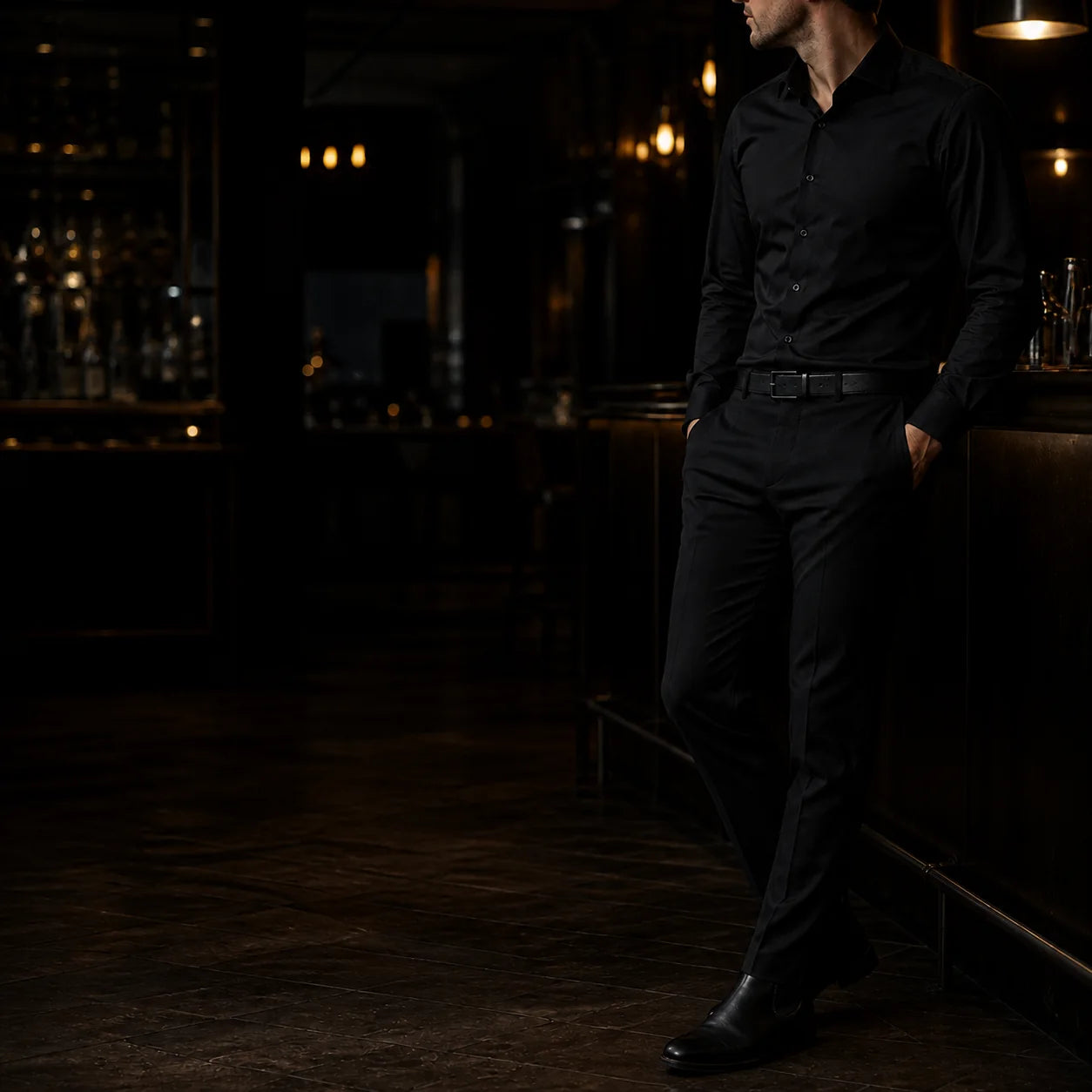 Man in an all-black shirt and trousers standing at a dimly lit bar, low three-quarter angle, face turned away from camera