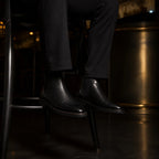 Black leather Chelsea boots resting on a barstool footrest, black trouser hem breaking over the boot