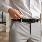 Close crop of a hand tucking the white shirt into light grey trousers above a black leather belt with a matte square buckle