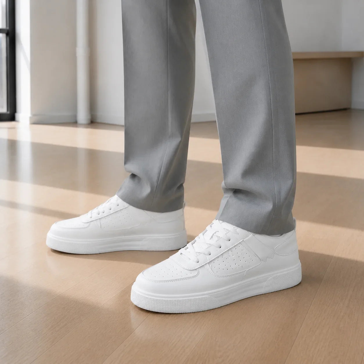 Man from the knee down on a light wood floor: plain white leather sneakers with a platform sole, the light grey trouser hem breaking cleanly over them