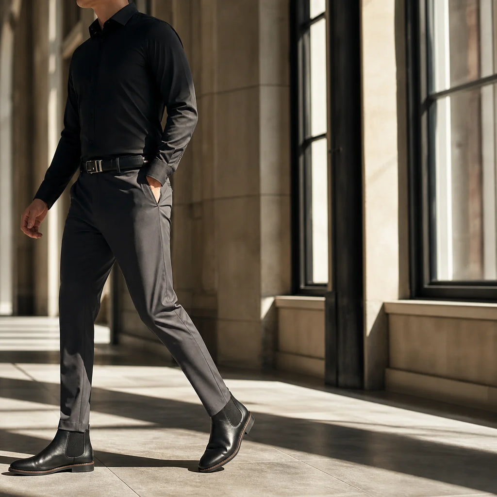 Man in a black shirt and charcoal trousers walking through a stone corridor in hard window light, head cropped above the chin