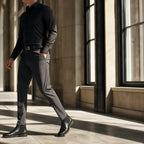 Man in a black shirt and charcoal trousers walking through a stone corridor in hard window light, head cropped above the chin