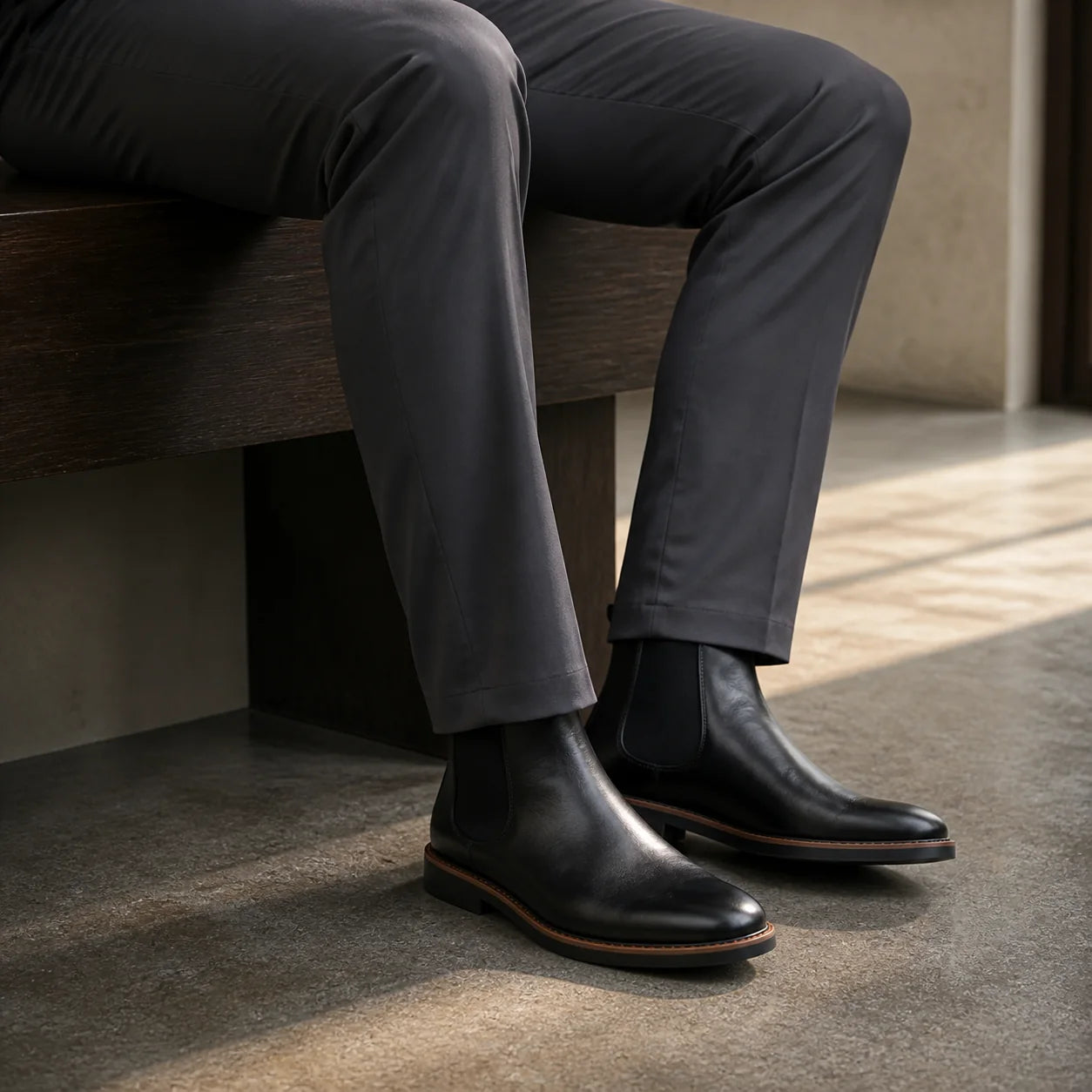 Seated man from the knee down: black leather Chelsea boots on stone floor, charcoal trouser hem breaking over the boot