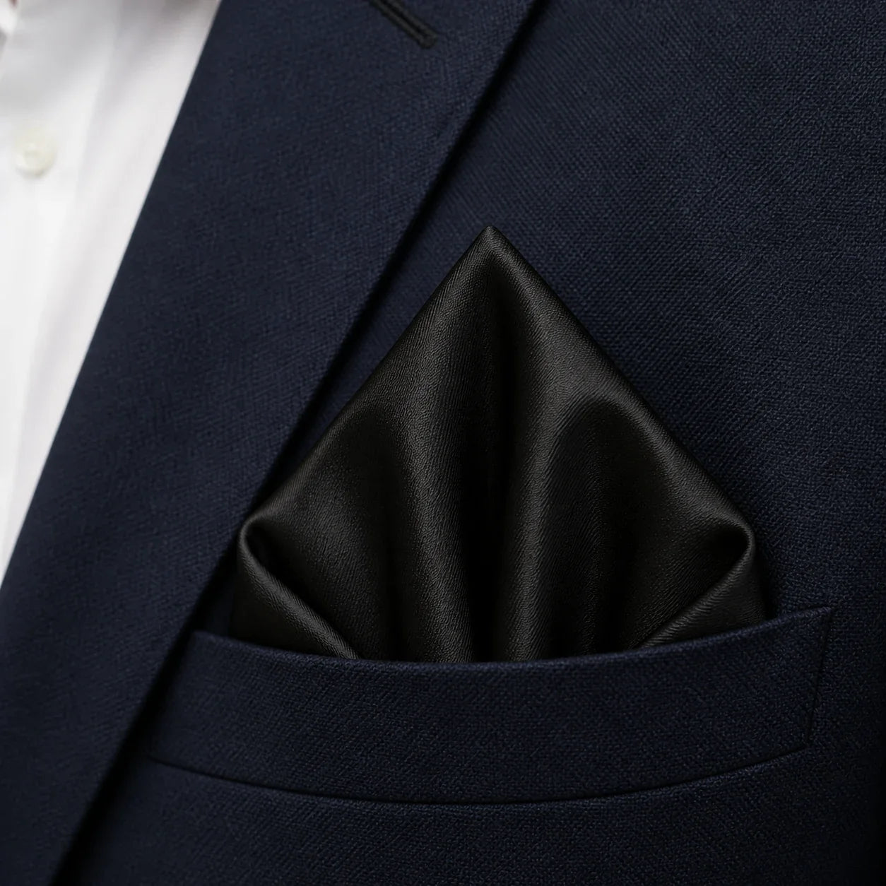 Close macro of the solid black pocket square folded into the breast pocket of the navy blazer, worn over a white shirt